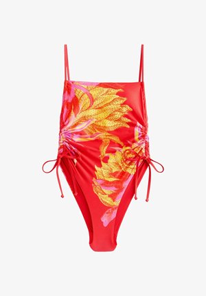 Kvinde i en rød og gul blomstret badedragt, der justerer sideremmen udendørs med grønne blade i baggrunden.