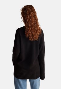 Czarny sweter dziany z prążkowanym dołem i teksturowanymi rękawami, o luźnym fasonie. Na plecach widać luźne loki czerwonych włosów.