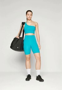 O conjunto de desporto em turquesa inclui um top de um ombro e calções ajustados; combinado com uma grande mala preta e tênis pretos.