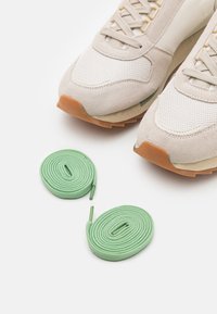 Sneaker beige e bianche con materiali in mesh e suede, caratterizzate da dettagli verdi, abbinate a due lacci rotondi di colore verde menta.