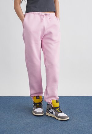 Osoba nosząca jasnoróżowe spodnie dresowe, z rękami w kieszeniach oraz szare, białe i żółte wysokie trampki, stojąca na niebieskim dywanie.