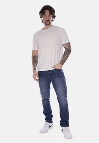 T-shirt di cotone bianco, jeans di denim blu e sneakers bianche. Presenta tatuaggi sulle braccia. Modello in piedi con le mani in tasca su uno sfondo semplice.