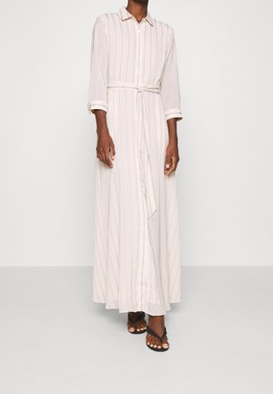 Femme portant une robe chemise longue rayée beige clair avec ceinture à nouer, boutonnée sur le devant, manches trois-quarts et sandales noires à bout ouvert.