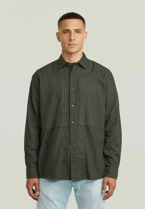 Ung mand med kort hår iført en mørkegrøn skjorte med knapper og lyseblå jeans, stående mod en ensartet lys baggrund.