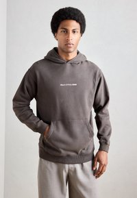Bruine hoodie met een voorkant kangaroo pocket en geribde manchetten. Bevat witte tekst "Marc O'Polo DENIM" op de borst. Gestyled met grijze broek.