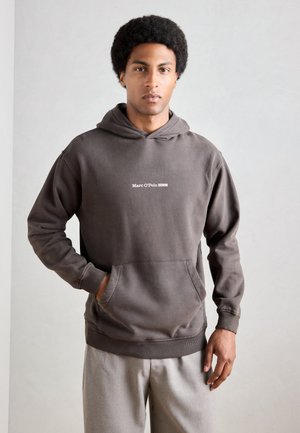 Brown hoodie with a front kangaroo pocket and ribbed cuffs. Features white text "Marc O'Polo DENIM" on the chest. Styled with gray pants.