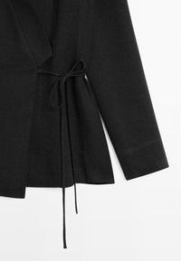 Haut portefeuille noir à manches longues avec fermeture nouée sur le côté, fabriqué en tissu texturé, montrant le détail de la manche et du nœud sur fond blanc.