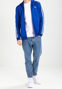 Blauwe sportjack met witte strepen op de mouwen, gedragen over een wit t-shirt. Gecombineerd met lichtblauwe jeans en witte sneakers.