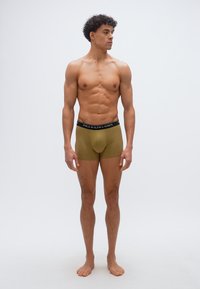 Άνδρας μοντέλο με μυώδη σωματότυπο, φορώντας χακί μπόξερ Ralph Lauren Polo, όρθιος ξυπόλητος μπροστά σε απλό ανοιχτό γκρι φόντο.