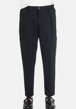 Pantaloni neri sartoriali con pieghe, indossati con scarpe nere in pelle intrecciata con fibbia, mostrati su una persona su uno sfondo semplice.