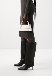 Sac à main en cuir blanc avec une poignée ronde, tenu dans une main, assorti à des bottes stiletto en cuir noir montant jusqu'aux genoux, sur une robe à motifs côtelés.