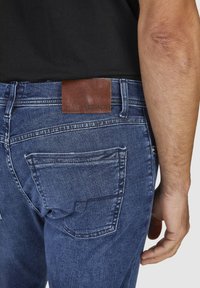 Granatowe dżinsy z widocznymi przeszyciami, pięcioma kieszeniami oraz brązową skórzaną naszywką z napisem "HECHNER" na tylnej stronie pasa.