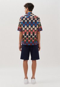 Kurzarmhemd mit buntem geometrischem Muster, mit Kragen und yoke auf der Rückseite, kombiniert mit navyblauen Shorts und weißen Sneakers.