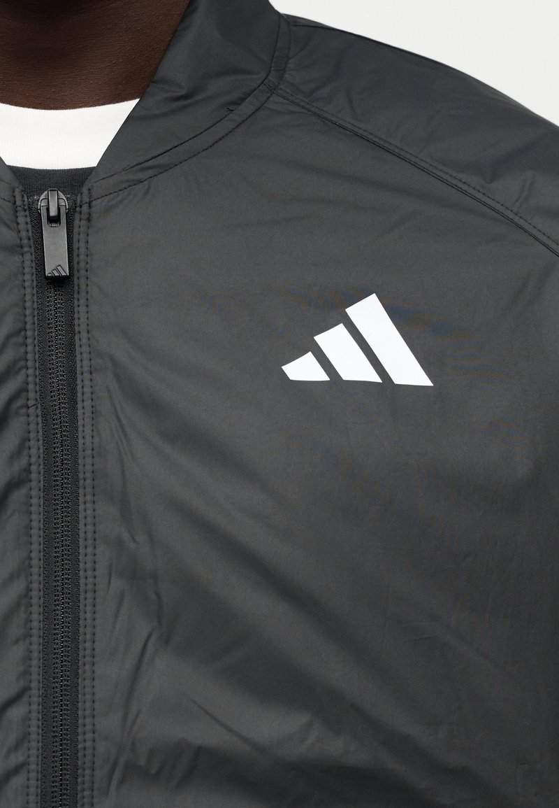Schwarze Windbreaker-Jacke mit durchgehendem Frontreißverschluss und weißem Drei-Streifen-Logo auf der linken Brust. Glattes, leichtes Material.