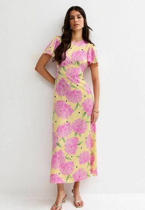 Donna con abito midi giallo con stampa floreale rosa e puntini neri, in piedi con un braccio sui fianchi su sfondo bianco.