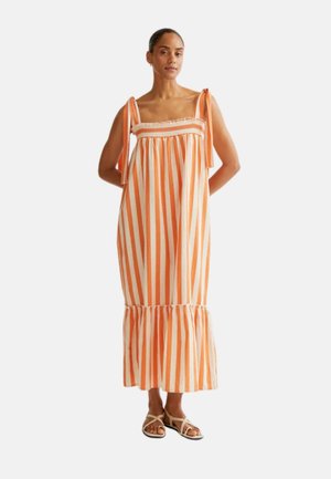 Mujer con vestido midi sin mangas de rayas verticales naranjas y blancas, con tirantes anudados en los hombros y dobladillo con volantes, combinado con sandalias blancas.