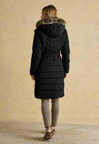 Naine, kes kannab musta vööga tikitud jopega, millel on karvane kapuuts, mustrilisi pükse ja pruune spordijalatsid, seismas beeži tekstuuriga seina vastu.