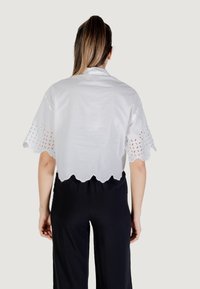 Camicetta bianca corta con motivi floreali ricamati sulle maniche e orlo scallop. Indossata con pantaloni neri, che mostrano uno stile casual.