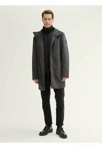 Grå yllemixkappa med ett innerlager med dragkedja, ståkrage och fickor. Bärs med svarta byxor och skor, mot en neutral bakgrund.