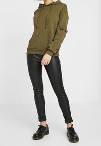 Sudadera con capucha verde oliva con un bolsillo tipo canguro, cordones y mangas con puños, combinada con leggings negros brillantes y zapatos negros con cordones.