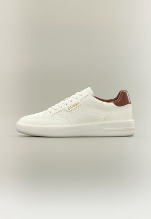 Zapatilla baja blanca con patrón en relieve, lengüeta trasera marrón, suela blanca y logotipo "GUESS" dorado en el lateral.