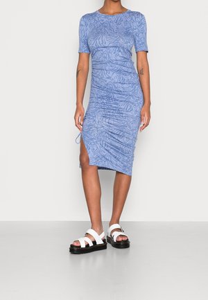 Kobieta ubrana w jasnoniebieską, krótkorękawową, marszczoną sukienkę midi z abstrakcyjnymi wzorami oraz białe sandały na platformie na jednolitym tle.