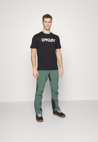 Sort t-shirt med hvid "Oakley" logo, parret med grønne og sorte bukser. Model iført sorte sko med hvide snørebånd, stående mod en ensfarvet baggrund.