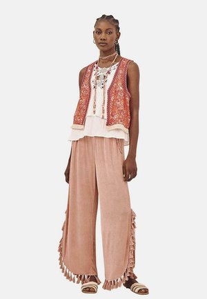 Conjunto en capas que incluye un chaleco rojo estampado sobre una blusa blanca, combinado con pantalones de pierna ancha de color rosa suave con detalles de flecos y sandalias.
