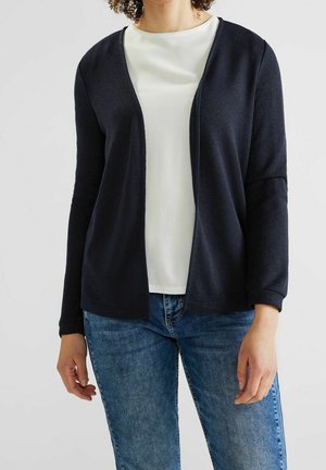 Femme portant un cardigan bleu marine ouvert sur un top blanc, associé à un jean bleu, sur un fond uni.