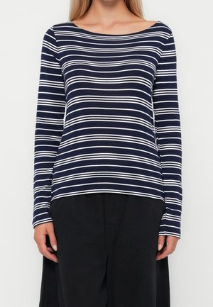 Femme portant une chemise à manches longues rayée bleu marine et blanc avec un pantalon noir, debout devant un fond blanc uni.