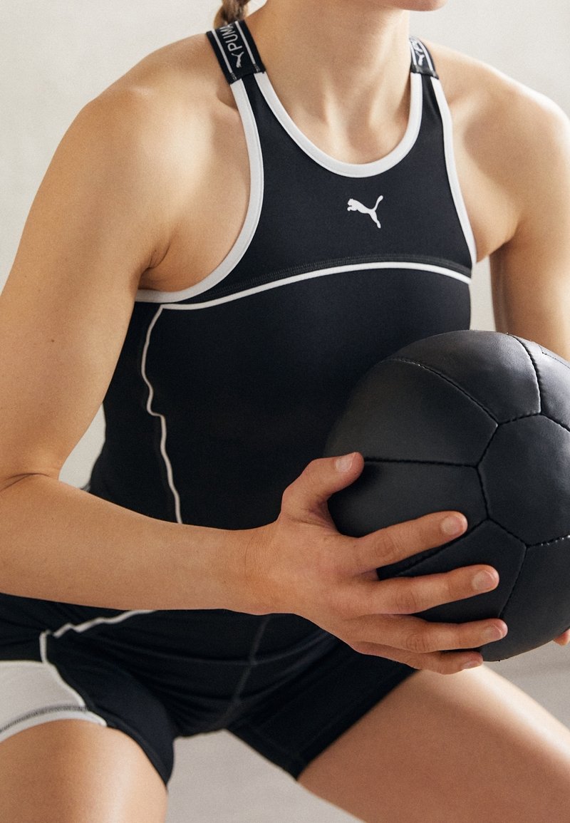 Svart atletisk linne med vita accenter och axelband, kombinerat med svarta shorts; håller en svart texturerad träningsboll.