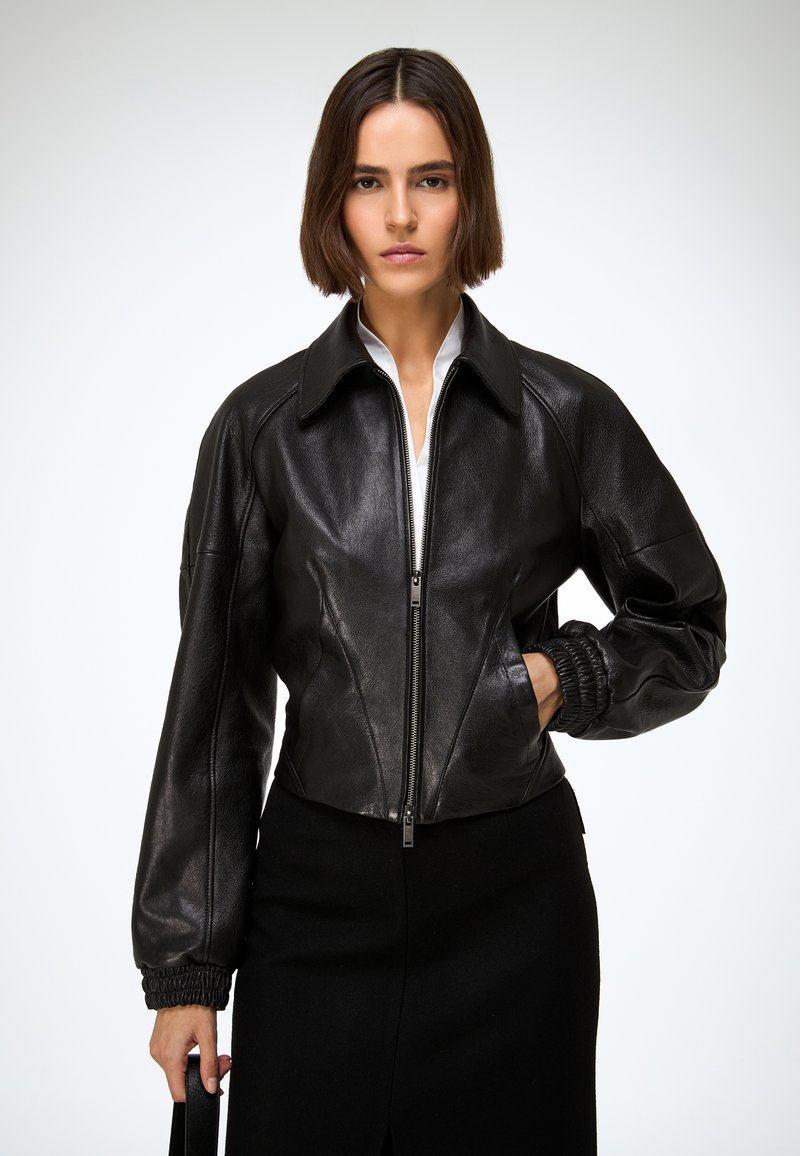 Crna kožna bomber jakna sa patentnim zatvaračem sprijeda, visokim ovratnikom i skraćenim krojem. Ima skupljene manžete i glatku teksturu.