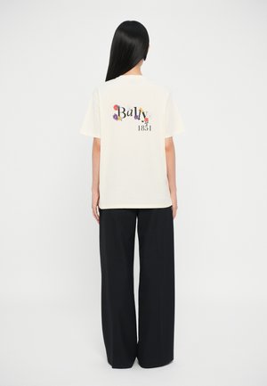 T-shirt in cotone crema con grafica floreale colorata e il testo "Bally 1851" sul retro. Abbinato a pantaloni neri a gamba larga.