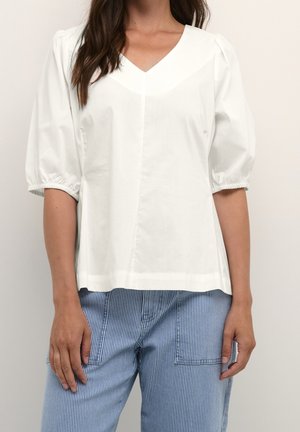 Kobieta ubrana w białą bluzkę z dekoltem w serek i bufkami oraz niebieskie spodnie w paski, stojąca na jednolitym tle.