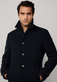 Uomo con capelli scuri che indossa un cappotto nero con bottoni e colletto rialzato, con le mani nelle tasche, che guarda direttamente nella telecamera su uno sfondo semplice.