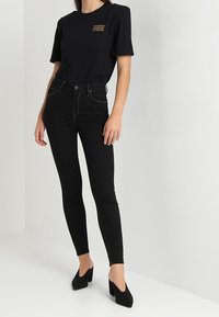 Černé bavlněné tričko s krátkým rukávem a zlatým grafickým textem, kombinované s černými úzkými džíny s vysokým pasem a černými sandály na podpatku.