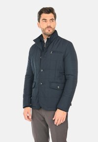 Giacca trapuntata blu navy con collo alto, bottoni frontali, due tasche laterali e una tasca sul petto con zip. Realizzata in materiale liscio e resistente all'acqua.