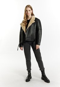 Giubbotto di pelle nera con colletto in shearling beige, design corto, chiusura con zip frontale, abbinato a pantaloni grigi slim-fit e stivaletti neri con tacco.
