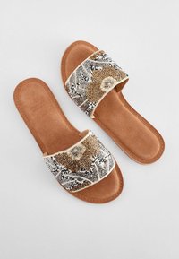 Flache Sandalen mit Perlenbesatz und einem breiten, dekorativen Riemen, der kunstvolle Muster in Gold, Silber und Schwarz auf einer braunen Wildledersohle zeigt.