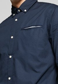 Mörkblå knapp-up skjorta i texturerad väv, med en liten ficka med vit accent och ett prickigt mönster genom hela skjortan.