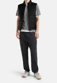 Man som bär en svart quiltad väst över en grå t-shirt, svarta byxor, vita sneakers, står med händerna vid sidorna mot en vit bakgrund.