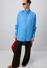 Nő oversized kék gombos inget visel, sötét széles szárú nadrágot, barna szandált, napszemüveget, piros táskát tart a kezében, és egy sima háttér előtt áll.