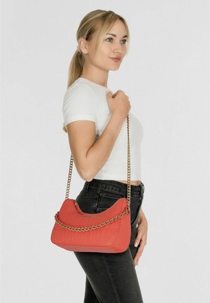 Sac à bandoulière en cuir rouge avec texture matelassée, bandoulière chaîne dorée, forme incurvée et logo embossé. Porté avec un jean noir.