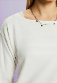 Biały bawełniany sweter o szerokim dekolcie i opadających ramionach, połączony z srebrnym naszyjnikiem z okrągłymi akcentami.