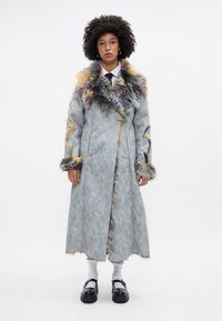 Garšs pelēks mētelis ar kažokādas akcentiem pelēkā un oranžā krāsā, ar augstu apkakli un atvērtu priekšpusi. Melni zābaki ar siksnām.