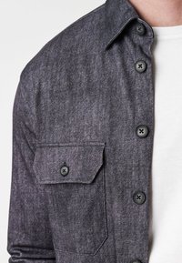 Button-up shirt van donkergrijze stof, met een patchzak met een knoop, een kraag en zes knopen aan de voorkant. Textured afwerking.