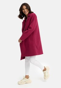 Fuchsia wollenblend lange jas met een notch-kraag, sluiting met één knoop en zijzakken, gedragen over witte broeken en beige sneakers.