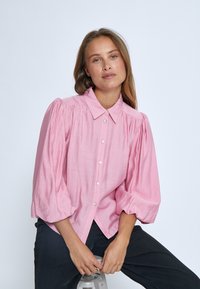 Blusa rosa con colletto e maniche a sbuffo, caratterizzata da bottoni sul davanti e un tessuto strutturato. Abbinata a pantaloni scuri.