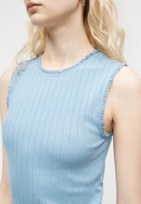 Γυναίκα με ξανθά μαλλιά που φοράει γαλάζιο αμάνικο ριμπ τοπ με διακριτικές βολάν στις άκρες, εμφανιζόμενη από τον λαιμό έως τα μέσα του κορμού.