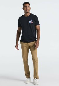 Man in een zwart T-shirt met patches, beige broek en witte sneakers, staande tegen een effen witte achtergrond.